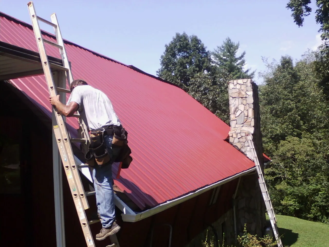 Roofer on a ladder beside a red metal roof and stone chimney — Soffit & Fascia Repair by Roofing Brothers in South Charleston